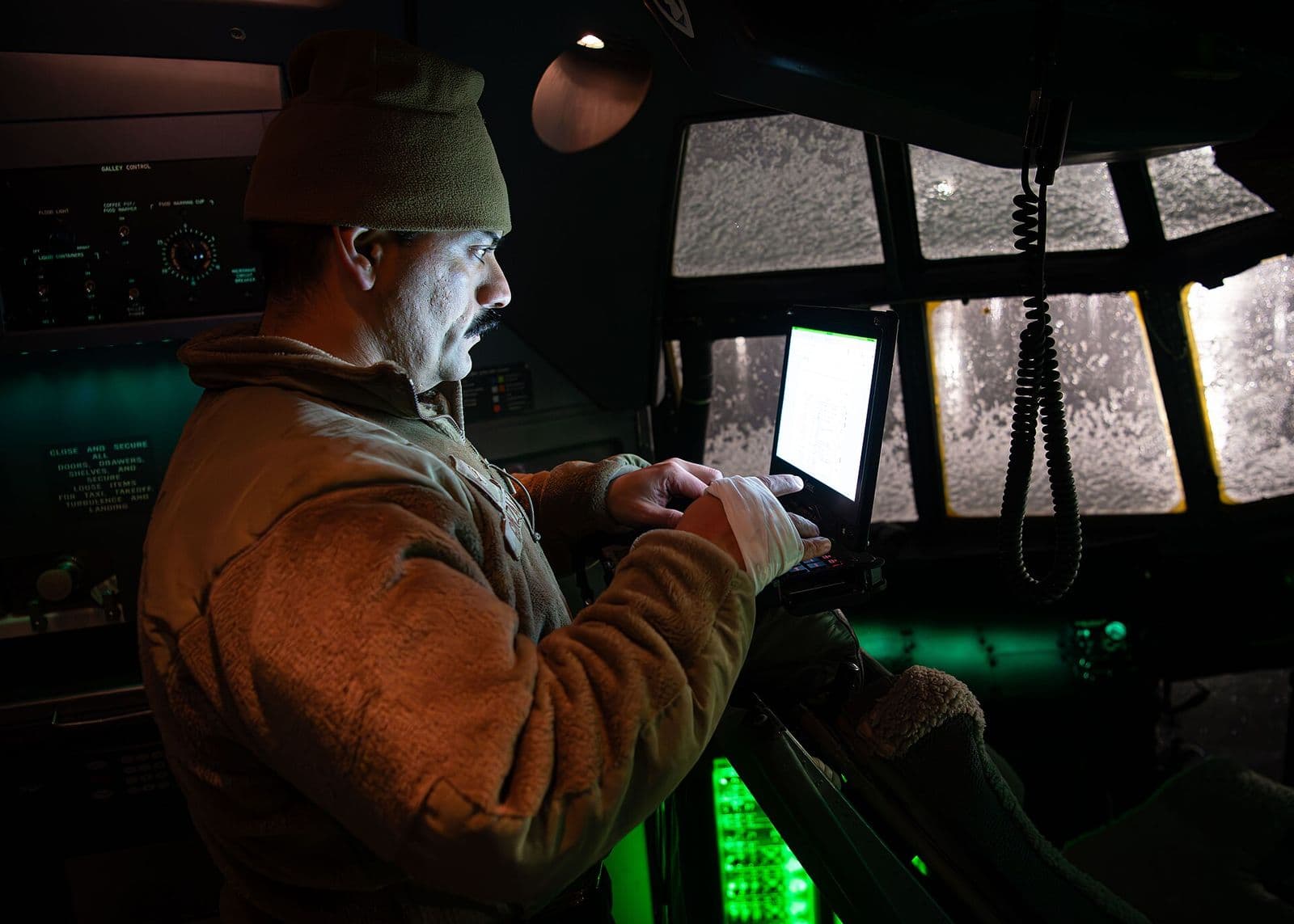 An avionics technician runs diagnostics on a laptop in the cockpit of a C-130J at night, instruments lit green