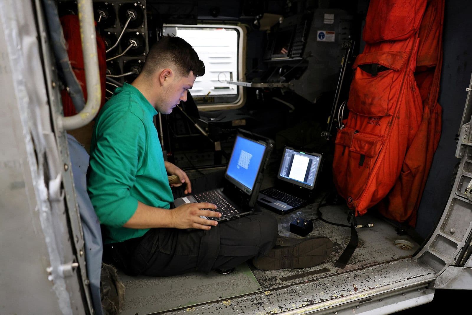 A sailor seated in a helicopter cabin records maintenance on a rugged laptop